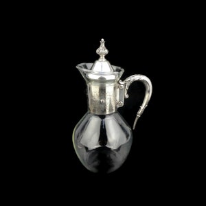May include: A silver-plated glass pitcher with a decorative handle and a silver lid. The pitcher has a clear glass body and a silver-plated base.