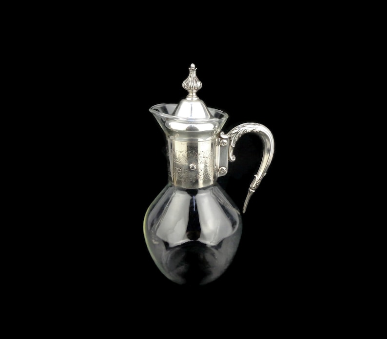 May include: A silver-plated glass pitcher with a decorative handle and a silver lid. The pitcher has a clear glass body and a silver-plated base.