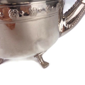 Puede incluir: Tetera de plata con un dise&ntilde;o floral decorativo alrededor del borde y tres patas. La tetera tiene un pico y una manija.