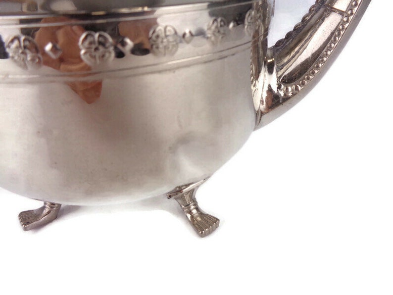 Puede incluir: Tetera de plata con un dise&ntilde;o floral decorativo alrededor del borde y tres patas. La tetera tiene un pico y una manija.