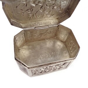 May include: A silver octagonal box with an intricate floral design on the lid and base. The box is open, revealing a plain interior.