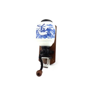 May include: A vintage wall-mounted coffee grinder with a white ceramic jar featuring a blue and white windmill scene. The grinder has a black metal crank and a wooden base.