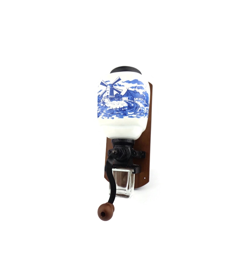 May include: A vintage wall-mounted coffee grinder with a white ceramic jar featuring a blue and white windmill scene. The grinder has a black metal crank and a wooden base.