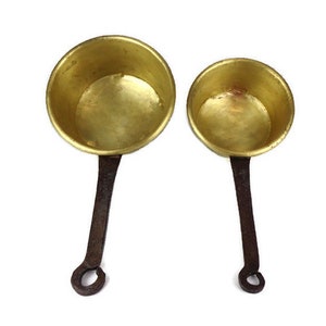 May include: Two brass ladles with long, dark iron handles. The ladles are different sizes, with the larger one in the background and the smaller one in the foreground.