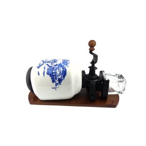 May include: A vintage coffee grinder with a white ceramic jar decorated with a blue and white windmill scene. The grinder has a black metal crank and a clear glass container for collecting coffee grounds.