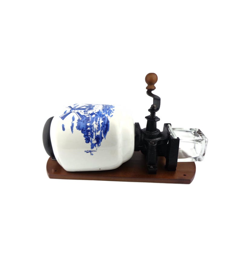 May include: A vintage coffee grinder with a white ceramic jar decorated with a blue and white windmill scene. The grinder has a black metal crank and a clear glass container for collecting coffee grounds.