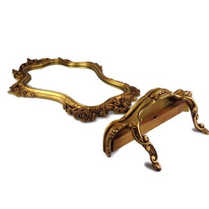 May include: A gold-toned ornate mirror with a decorative frame and a stand. The mirror is oval-shaped and has a scalloped edge. The stand is made of wood and has a decorative design.