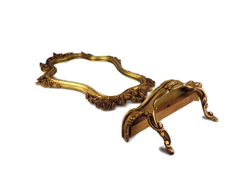 May include: A gold-toned ornate mirror with a decorative frame and a stand. The mirror is oval-shaped and has a scalloped edge. The stand is made of wood and has a decorative design.