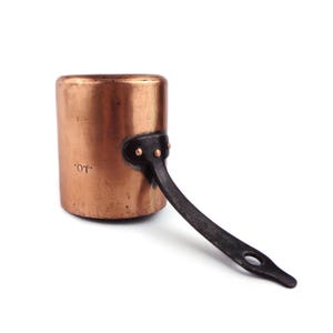 May include: A small copper saucepan with a dark metal handle. The copper pot has a cylindrical shape and is marked with the initials "J.O." The handle is attached with rivets. The pot is likely vintage or antique.