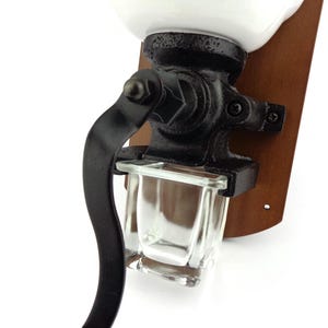 May include: A vintage-style, wall-mounted coffee grinder with a white ceramic top, black metal crank, and a glass collection bin. The grinder is mounted on a wooden base.
