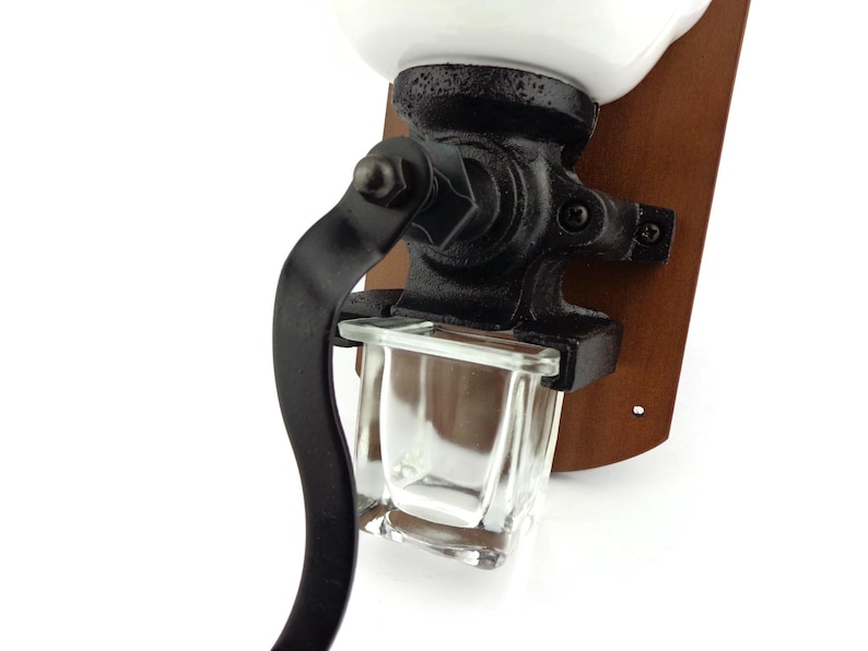 May include: A vintage-style, wall-mounted coffee grinder with a white ceramic top, black metal crank, and a glass collection bin. The grinder is mounted on a wooden base.