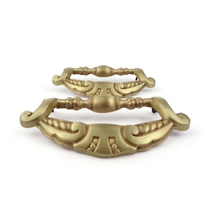 May include: Three antique brass drawer pulls with ornate, symmetrical designs. The pulls have a curved shape with decorative detailing, including a central handle and stylized end pieces. The brass finish gives them a vintage look.