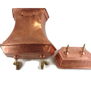 May include: A copper planter with a square base and a flared top, featuring four decorative feet. The planter has a separate lid with two decorative knobs.