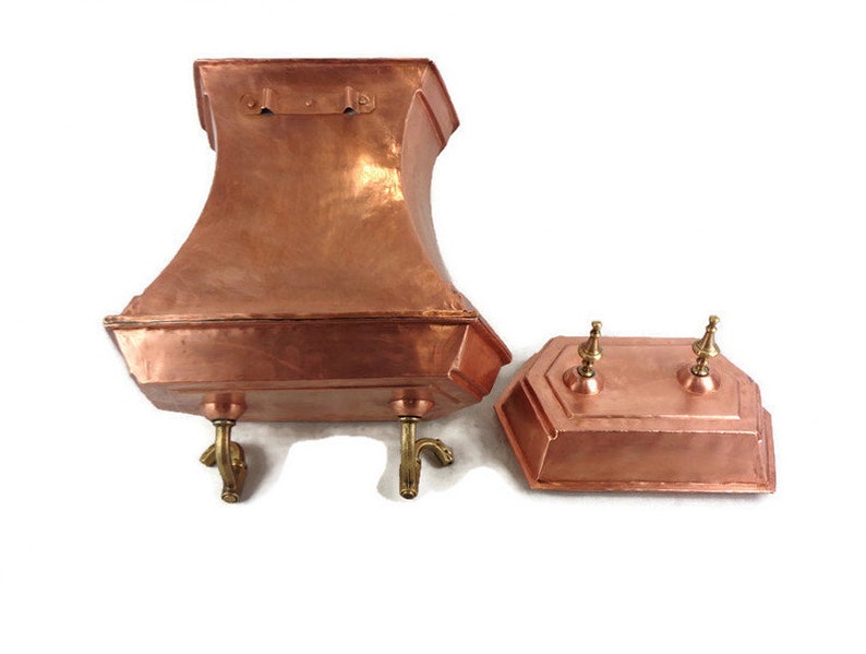 May include: A copper planter with a square base and a flared top, featuring four decorative feet. The planter has a separate lid with two decorative knobs.