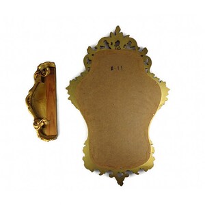 May include: A gold-colored ornate mirror with a decorative frame. The mirror is oval-shaped and has a brown backing. The frame has a detailed design with intricate carvings. The mirror is hanging on a wall.