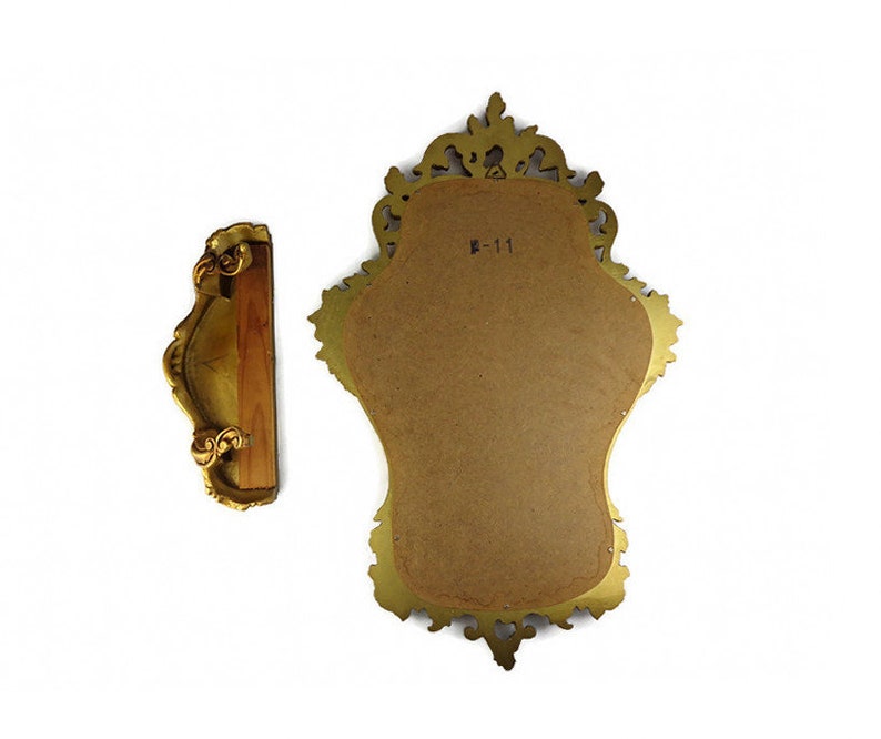 May include: A gold-colored ornate mirror with a decorative frame. The mirror is oval-shaped and has a brown backing. The frame has a detailed design with intricate carvings. The mirror is hanging on a wall.