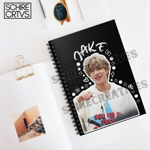 May include: Black spiral notebook with a white border and the name "JAKE" written in white. A photo of a young man with brown hair is on the cover. The photo is surrounded by white doodles.