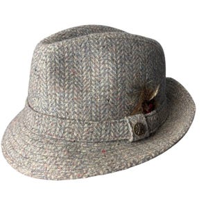 May include: A gray tweed fedora hat with a herringbone pattern. The hat features a decorative feather and a small, round button on the band. The brim is slightly upturned, and the hat has a classic, vintage style.