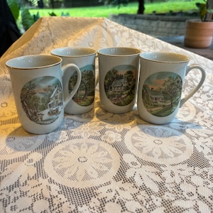 May include: Four white ceramic mugs with gold rims and handles. Each mug features a circular landscape scene with a house, trees, and a sky. The mugs are arranged on a white lace tablecloth.