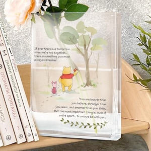 以下が含まれることがあります： 透明な長方形の花瓶。くまのプーさんのイラストとインスピレーションを与える引用句が描かれています。花瓶には白と桃色の花が入っています。左側にはトルコ語の本が並んでいます。