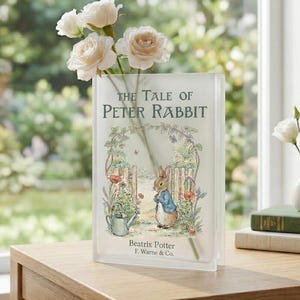 May include: A clear glass vase shaped like a book, featuring the cover art of "The Tale of Peter Rabbit" by Beatrix Potter. The vase holds white roses. The scene is set on a wooden table near a window.