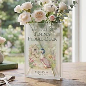 May include: A clear glass vase shaped like a book, filled with white roses, lavender, and eucalyptus. The vase features the cover art of "The Tale of Jemima Puddle-Duck" by Beatrix Potter. A small vase with flowers and a book are on a wooden table.