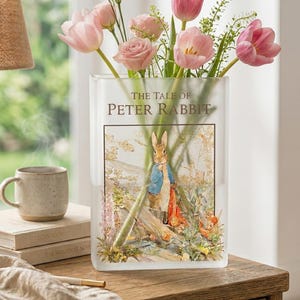May include: A clear glass vase with a floral arrangement of pink tulips and roses. The vase features an illustration from "The Tale of Peter Rabbit." A mug and books sit on a wooden table next to the vase.