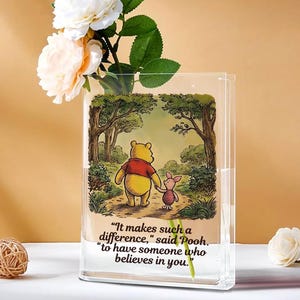 Puede incluir: Jarrón rectangular transparente con una ilustración de Winnie the Pooh. La imagen muestra a Pooh y Piglet en un bosque. El jarrón contiene rosas blancas y melocotón. El texto en el jarrón dice: "Marca la diferencia, dijo Pooh, tener a alguien que crea en ti."