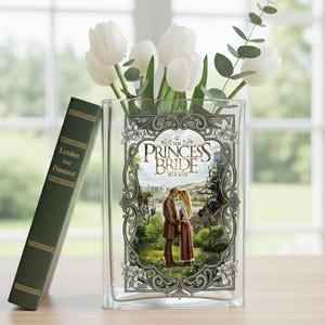 May include: A clear glass vase featuring an image from "The Princess Bride", filled with white tulips and foliage. A green book with gold lettering rests beside it. The vase is on a wooden surface.