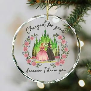 May include: A clear, faceted glass ornament with a gold string. The ornament features a fairytale scene with two figures in gowns, a castle, and the text "Changed for good because I knew you."