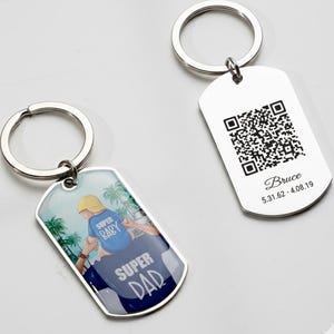 Puede incluir: Dos llaveros plateados. Uno presenta un gráfico de un niño sobre la espalda de una persona con las palabras "Super Baby" y "Super Dad". El otro llavero tiene un código QR y el nombre "Bruce" con fechas.
