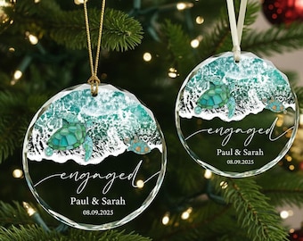 May include: Two clear, round Christmas ornaments with a sea turtle design. The ornaments have a turquoise and white wave pattern at the top, with the word "engaged" and the names "Paul & Sarah" and the date 08.09.2025.