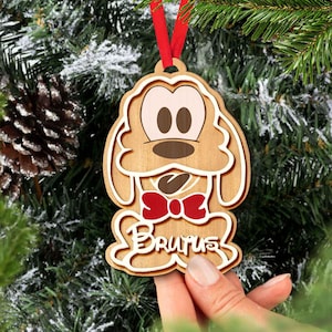 May include: Wooden Christmas bauble shaped like a cartoon dog with brown fur, white eyes, and a red bow tie. The bauble has the name "Brutus" written on it.