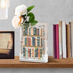 May include: A clear, rectangular vase with a bookshelf design, filled with white and peach-colored flowers. The vase is decorated with colorful books and floral accents. A framed picture and a stack of books are also visible.