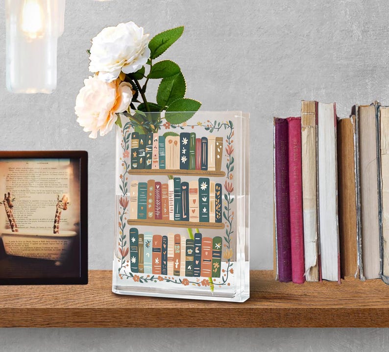 May include: A clear, rectangular vase with a bookshelf design, filled with white and peach-colored flowers. The vase is decorated with colorful books and floral accents. A framed picture and a stack of books are also visible.