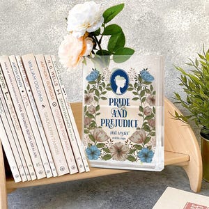 K&ouml;nnte beinhalten: Transparente Acryl-Spielkartenbox mit dem Titel "Pride and Prejudice" und floralen Designs. Ein Stapel B&uuml;cher und eine Vase mit Blumen sind ebenfalls zu sehen.