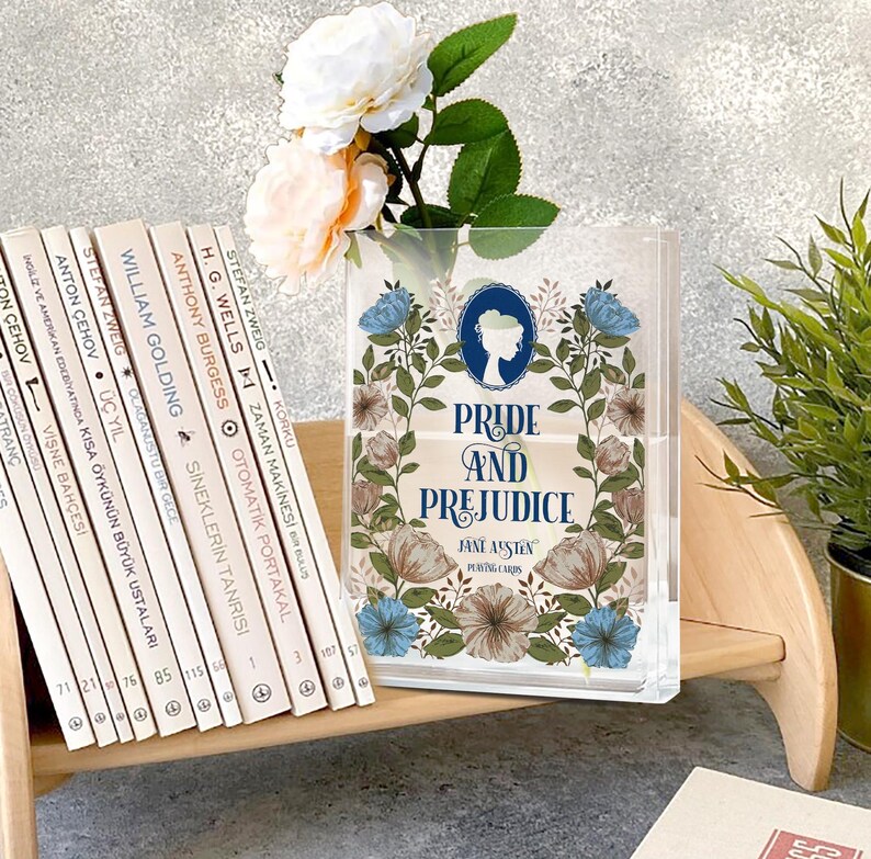 K&ouml;nnte beinhalten: Transparente Acryl-Spielkartenbox mit dem Titel "Pride and Prejudice" und floralen Designs. Ein Stapel B&uuml;cher und eine Vase mit Blumen sind ebenfalls zu sehen.