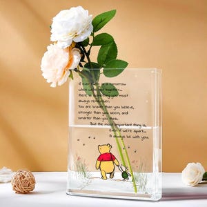 May include: A clear, rectangular glass vase with a Winnie the Pooh illustration and inspirational text. The vase holds white and peach-colored roses and green foliage. A decorative woven ball sits in the foreground.