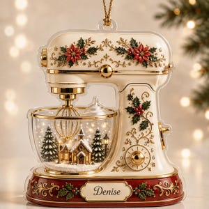 Könnte beinhalten: Ein cremefarbener Standmixer-Anhänger mit goldenen Akzenten, roten Weihnachtssternen und grünen Stechpalmenblättern. Die klare Rührschüssel enthält eine winterliche Miniatur-Dorfszene mit verschneiten Häusern. Ein Namensschild zeigt Denise.