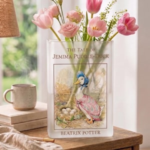 May include: A rectangular glass vase featuring a floral arrangement of pink tulips and roses. The vase is decorated with an illustration from "The Tale of Jemima Puddle-Duck" by Beatrix Potter. The vase sits on a wooden table.