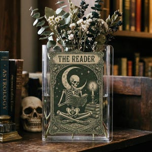 Peut inclure: Un vase en verre transparent avec un panneau décoratif représentant un squelette lisant sous un croissant de lune, avec le texte "THE READER". Le vase contient des fleurs séchées et est posé sur une surface en bois, avec des livres en arrière-plan.