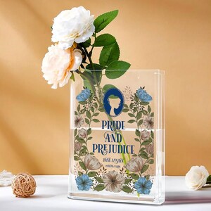 K&ouml;nnte beinhalten: Eine transparente, rechteckige Vase mit dem Text "Pride and Prejudice" und floralen Mustern. Die Vase enth&auml;lt wei&szlig;e und pfirsichfarbene Rosen und gr&uuml;nes Laub. Eine kleine dekorative Kugel befindet sich im Vordergrund.