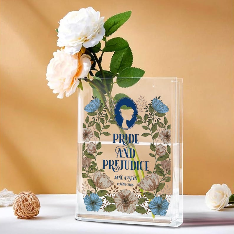 K&ouml;nnte beinhalten: Eine transparente, rechteckige Vase mit dem Text "Pride and Prejudice" und floralen Mustern. Die Vase enth&auml;lt wei&szlig;e und pfirsichfarbene Rosen und gr&uuml;nes Laub. Eine kleine dekorative Kugel befindet sich im Vordergrund.