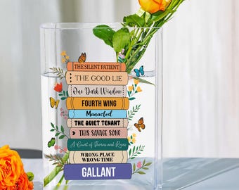 Puede incluir: Un jarrón de vidrio transparente con títulos de libros impresos, incluyendo "The Silent Patient" y "Gallant". El jarrón contiene agua y flores frescas, adornado con mariposas y motivos florales. El jarrón es rectangular.