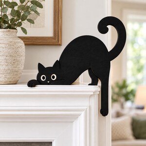 May include: A black cat silhouette figurine crafted from wood, featuring a curved tail and wide white eyes. The cat is positioned in a stretching pose with its front paws resting on the edge of a white mantelpiece and one back leg hanging down.