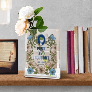 K&ouml;nnte beinhalten: Eine transparente, rechteckige Vase mit dem Text "Pride and Prejudice" und floralen Illustrationen. Die Vase enth&auml;lt wei&szlig;e und pfirsichfarbene Blumen. Ein gerahmtes Bild und eine Reihe von B&uuml;chern sind im Hintergrund zu sehen.