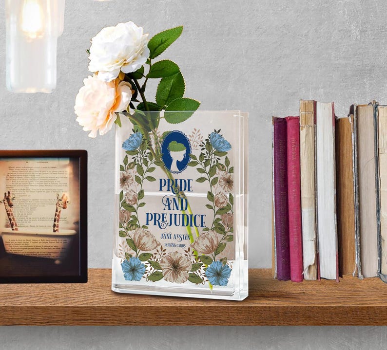K&ouml;nnte beinhalten: Eine transparente, rechteckige Vase mit dem Text "Pride and Prejudice" und floralen Illustrationen. Die Vase enth&auml;lt wei&szlig;e und pfirsichfarbene Blumen. Ein gerahmtes Bild und eine Reihe von B&uuml;chern sind im Hintergrund zu sehen.