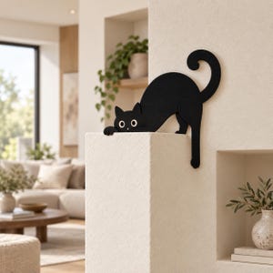 May include: A flat, black silhouette of a cat with large white eyes and a curled tail, designed to be mounted on the corner of a wall. The cat is depicted in a crouching position with its front paws extended over the edge of the wall surface.