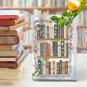 May include: A clear glass vase with a bookshelf design, filled with yellow and orange flowers. The vase has a floral border and is next to a stack of old books. The bookshelf design features colorful book spines.