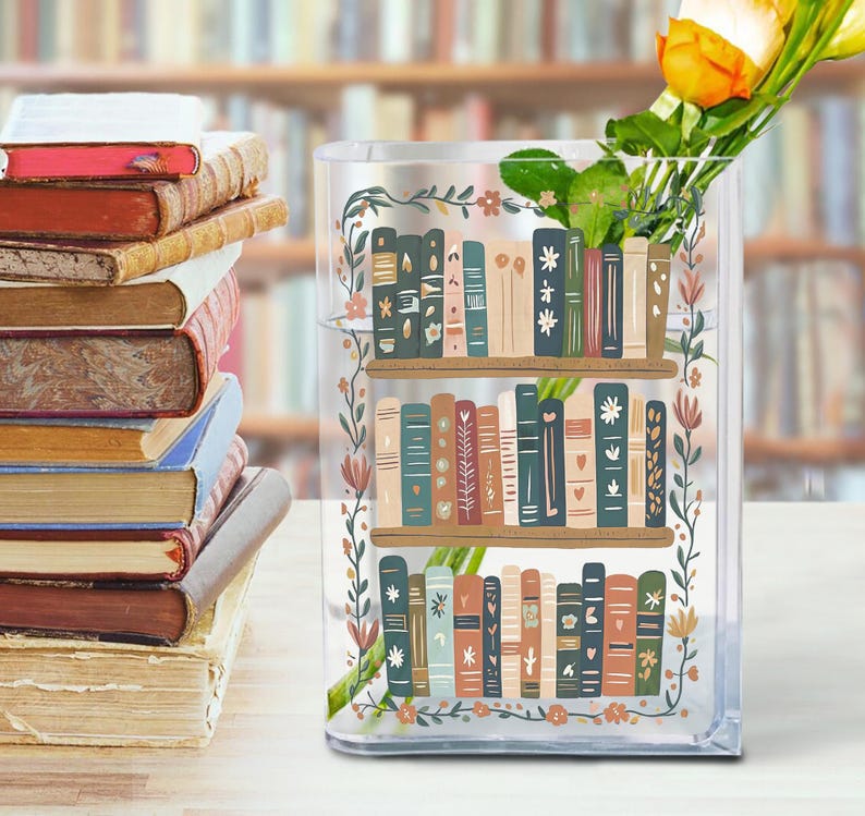 May include: A clear glass vase with a bookshelf design, filled with yellow and orange flowers. The vase has a floral border and is next to a stack of old books. The bookshelf design features colorful book spines.